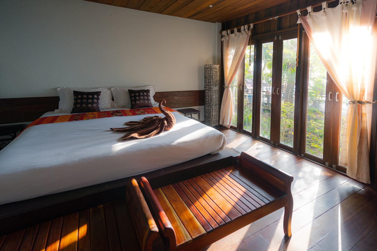 Sunlit wooden cabin bedroom with white bedding, towel folded into a swan shape, and glass doors opening to green trees